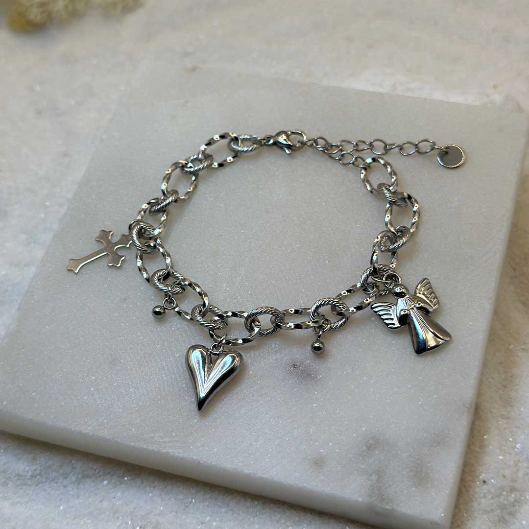 Un magnifique bracelet en acier inoxydable "Angely" est au centre de l'image. Le bijou possède des charmes : un ange, une croix et un coeur. Le bracelet est posé sur une plaque de marbre. Il y a du sable blanc en fond.