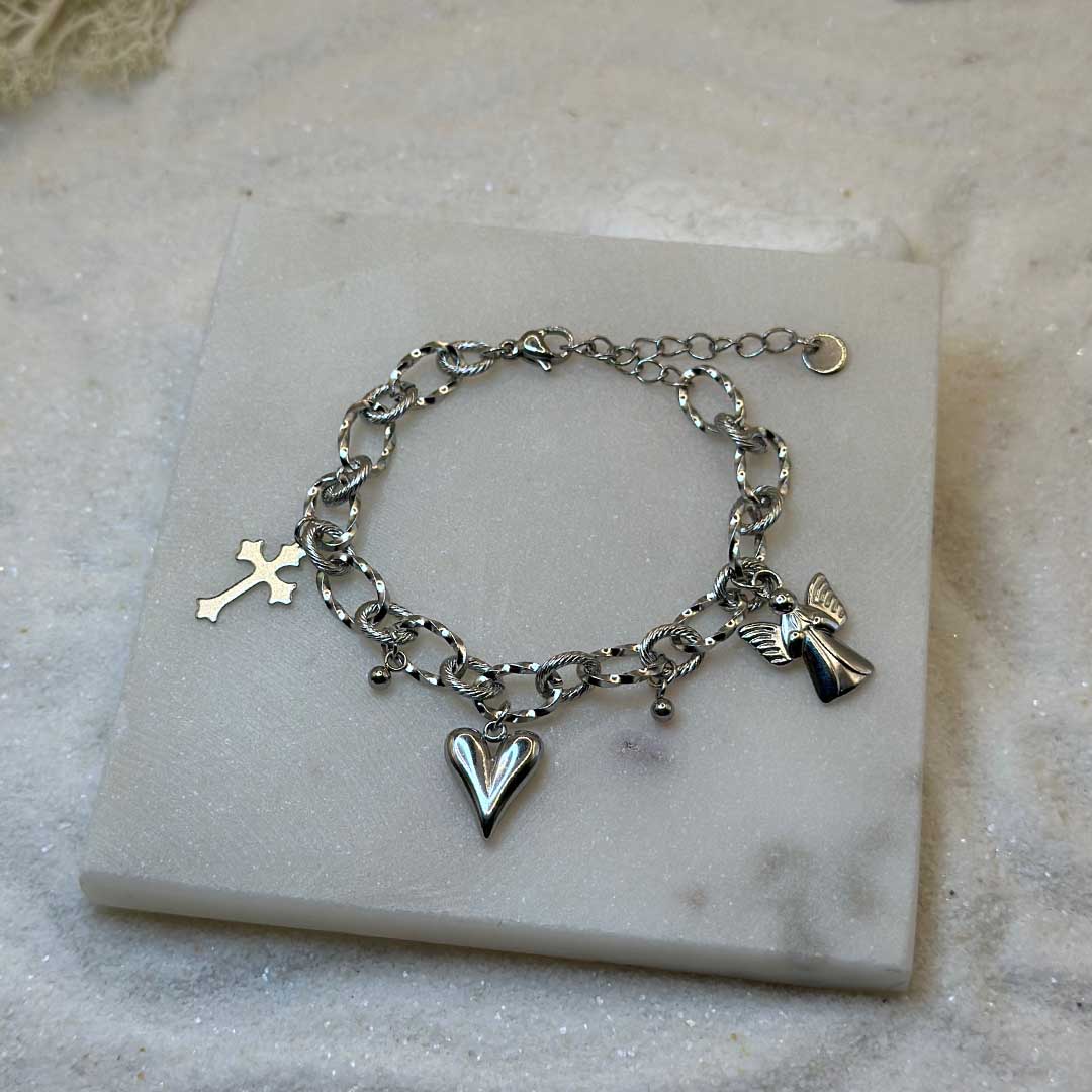 Un magnifique bracelet en acier inoxydable "Angely" est au centre de l'image. Le bijou possède des charmes : un ange, une croix et un coeur. Le bracelet est posé sur une plaque de marbre. Il y a du sable blanc en fond.