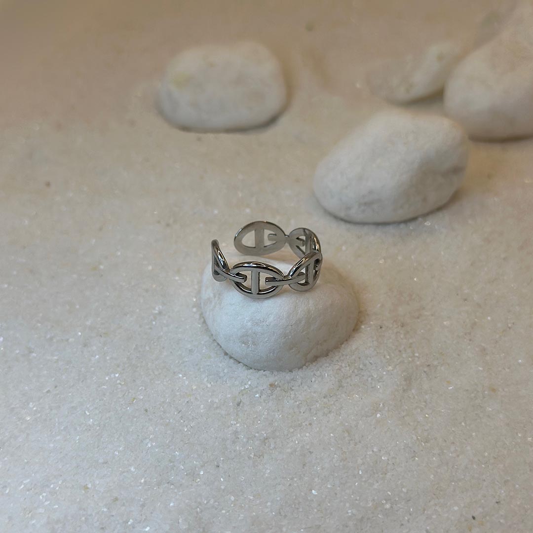 Une très jolie bague en argent "ari" est au centre de l'image, posée sur un caillou blanc. La bague est sur le dessus de la pierre. Le bijoux en acier inoxydable est en forme de maillons. Tout autour se trouve du sable blanc et d'autres cailloux.