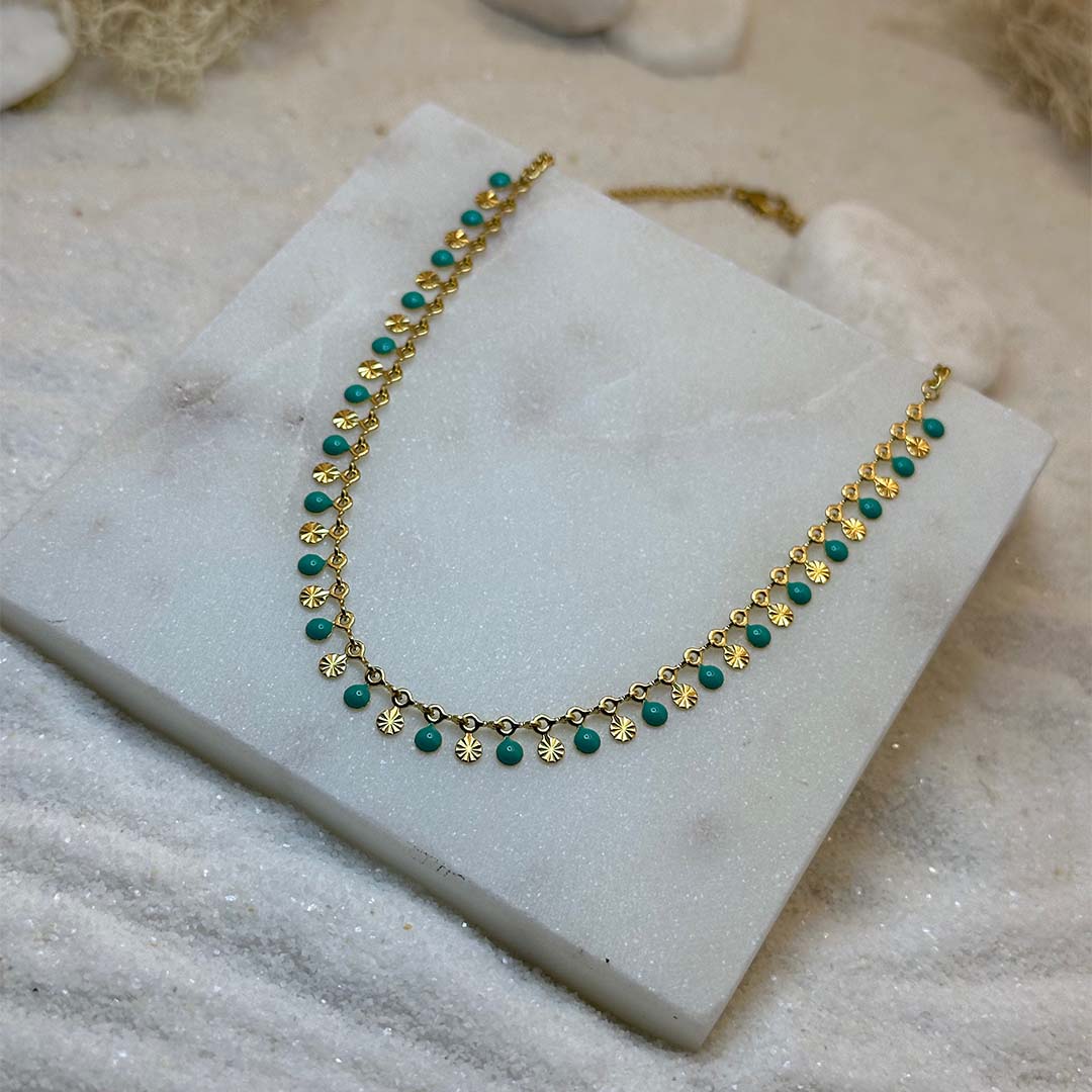 Un magnifique collier en or "ariella" est au centre de l'image, vu de 3/4, posé sur une grosse plaque de marbre. Le bijoux en acier inoxydable est composée d'une chaine en or, de charms en forme de coquillage et de petits rond verts. Autour se trouve du sable blanc.