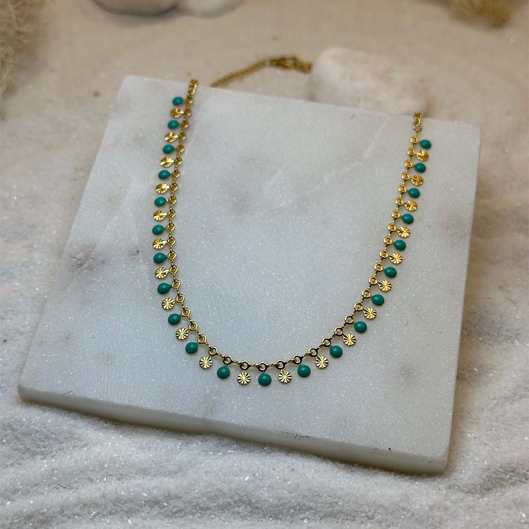 Un magnifique collier en or "ariella" est au centre de l'image, vu de face, posé sur une grosse plaque de marbre. Le bijoux en acier inoxydable est composée d'une chaine en or, de charms en forme de coquillage et de petits rond verts. Autour se trouve du sable blanc.