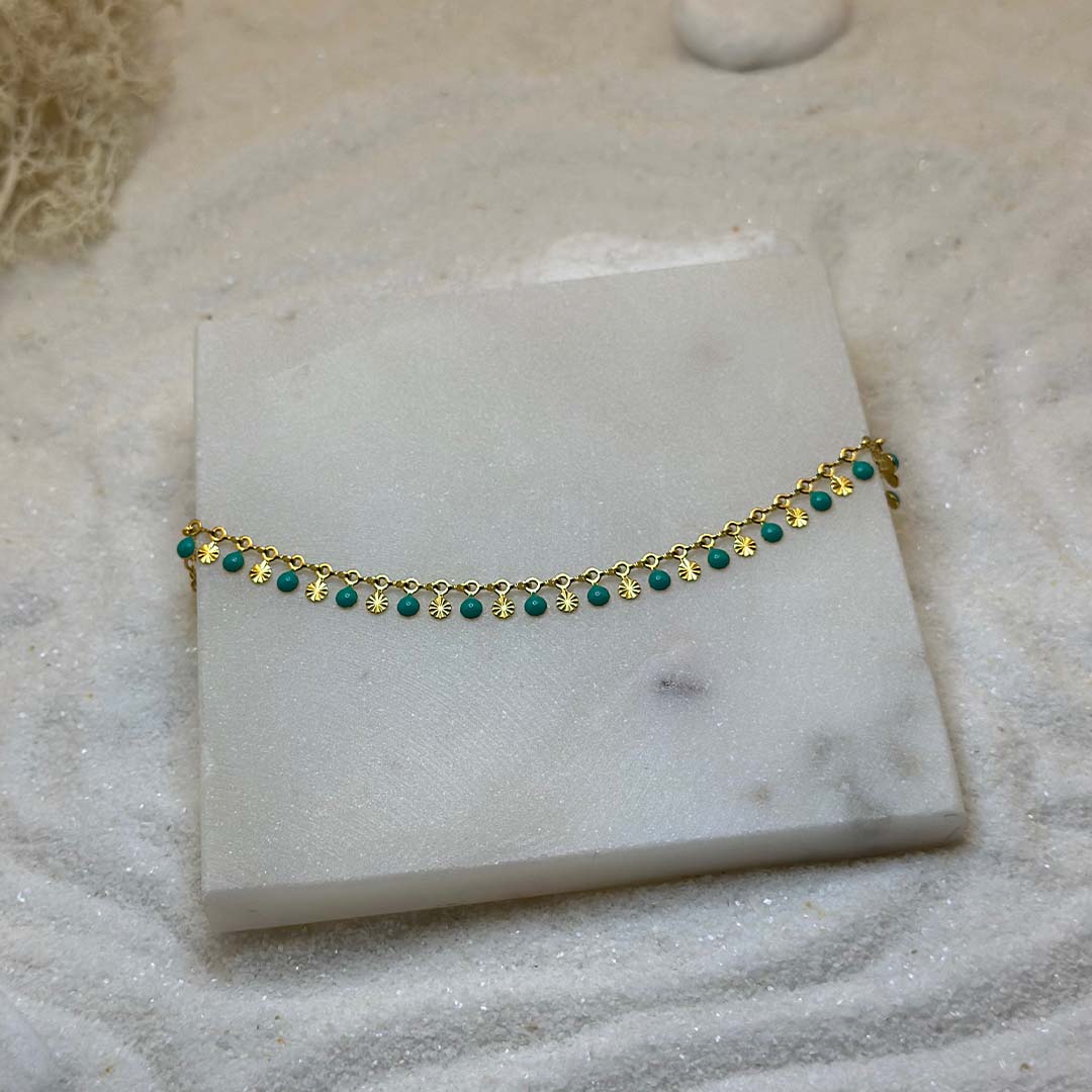 Un magnifique bracelet en or "ariella" est au centre de l'image, vu de 3/4, posé sur une grosse plaque de marbre. Le bijoux en acier inoxydable est composée d'une chaine en or, de charms en forme de coquillage et de petits rond verts. Autour se trouve du sable blanc.