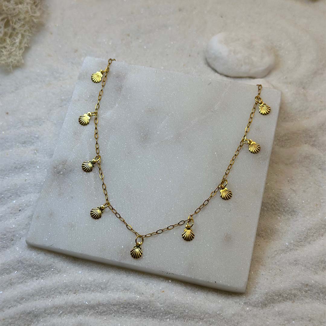 Un magnifique collier en acier inoxydable "Atia" de couleur or, ce collier possède des forme de coquillage or tout le long de la chaine, le bijou est posé sur un plaque de marbre blanchec qui est posé sur du sable blanc qui fait office de fond d'image.