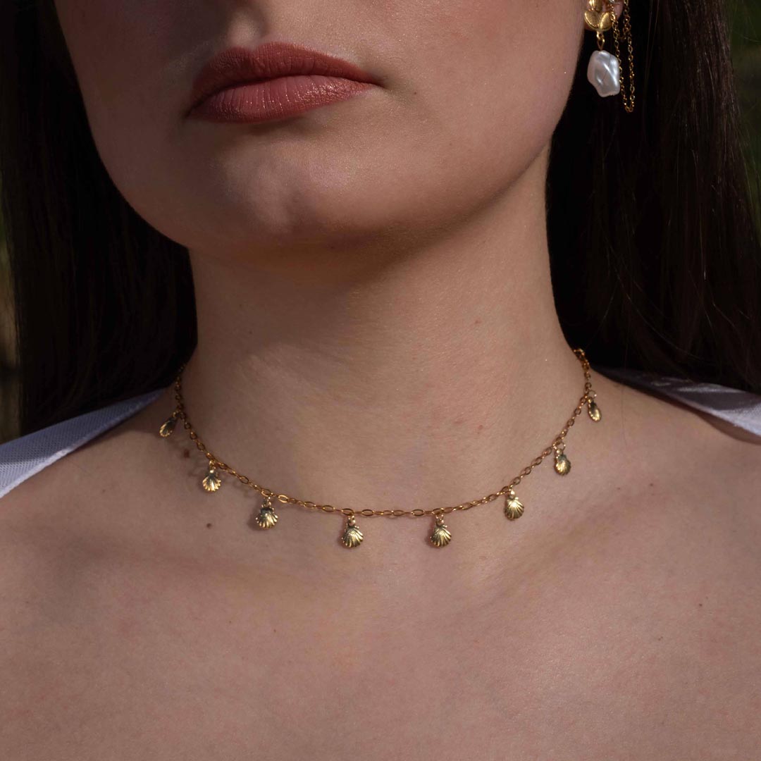 Photo d'un cou et d'un bas de visage montrant ce magnifique bijou "atia" en acier inoxydable qui est un collier avec des petits charms coquillages de couleur doré. Le bijou est de couleur doré / or.