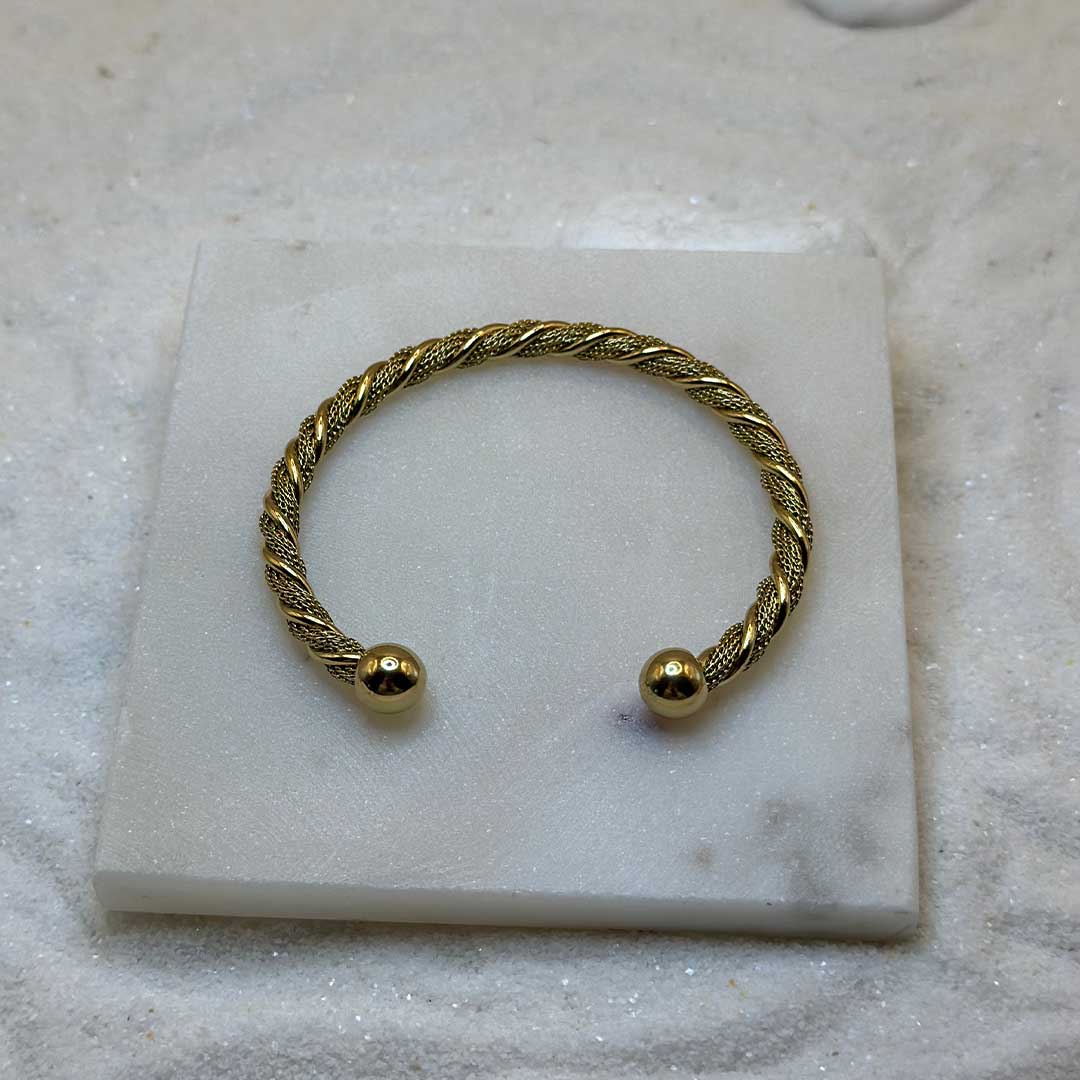 Un jolie bracelet en or "atilia" est au centre de l'image, posé sur une plaque de marbre. Le bijoux en acier inoxydable est moulé avec des reliefs et possède deux boules en or à chaque extrémités. Tout autour se trouve du sable blanc.