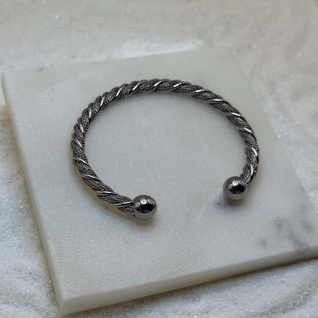 Un joli bracelet en argenté "atilia" est au centre de l'image, posé sur une plaque de marbre. Le bijoux en acier inoxydable est moulé avec des reliefs et possède deux boules en aregnté à chaque extrémités. Tout autour se trouve du sable blanc.