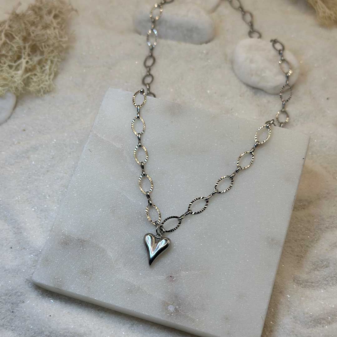 Un collier très mignon en argent "carla" est au centre de l'image, posé sur une plaque de marbre. Le bijoux en acier inoxydable est composé d'une chaîne de maillons en argent, ainsi que d'une pendentif en forme de coeur. Tout autour se trouve du sable blanc, des cailloux blanc et des algues.