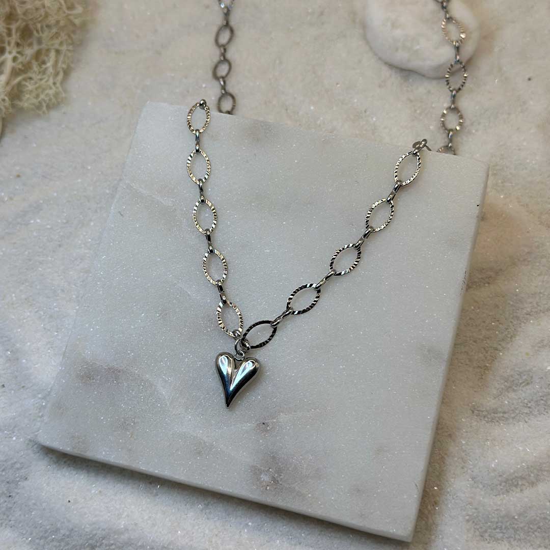 Un collier très mignon en argent "carla" est au centre de l'image en gros plan, posé sur une plaque de marbre. Le bijoux en acier inoxydable est composé d'une chaîne de maillons en argent, ainsi que d'une pendentif en forme de coeur. Tout autour se trouve du sable blanc, des cailloux blanc et des algues.