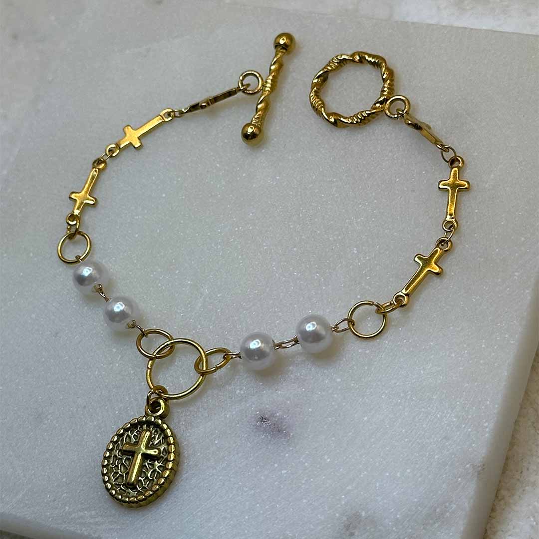 Un magnifique collier en or "charlie" est au centre de l'image en gros plan. C'est un très beau bracelet en acier inoxydable composé de perles, de croix, de maillons, ainsi que d'un pendentif ovale avec une croix. Le bijoux est posé sur une plaque de marbre. Tout autour se trouve du sable blanc.