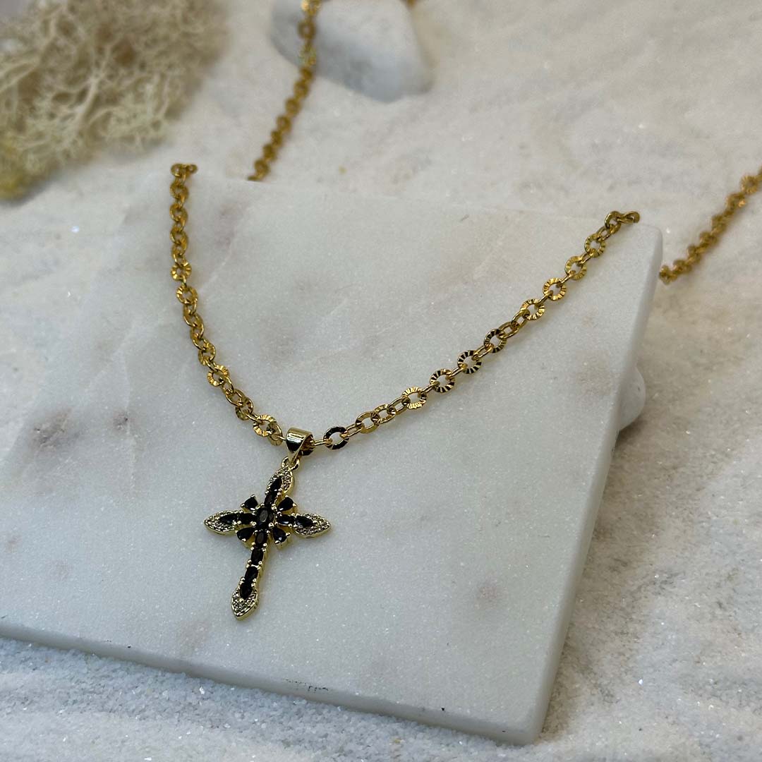 Un très jolie collier en or "crecia" est au centre de l'image, posé sur une plaque de marbre. Le bijoux en acier inoxydable est composé d'une chaîne en or et d'un pendentif en forme de croix. Tout autour se trouve du sable blanc.