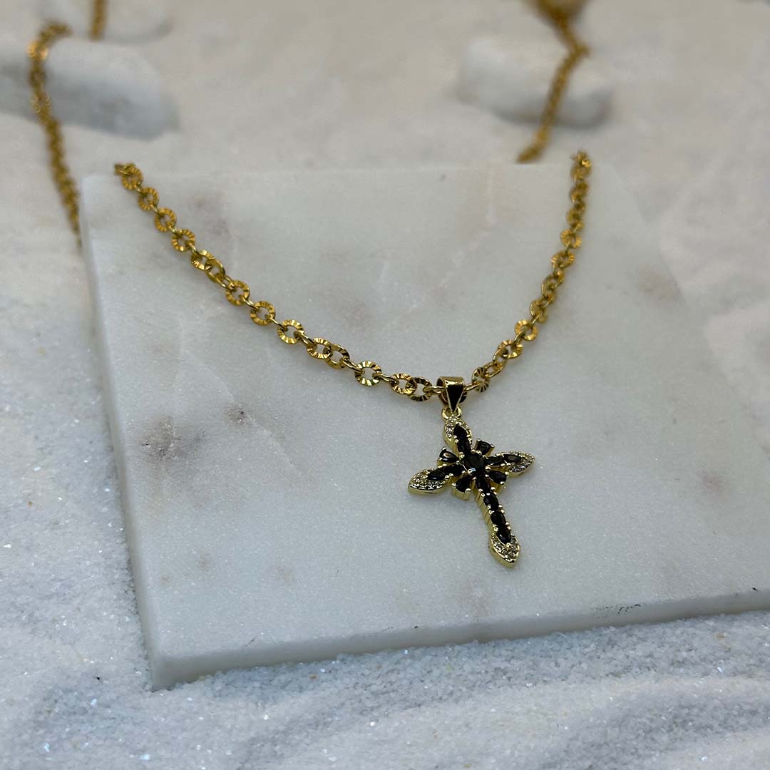 Un très jolie collier en or "crecia" est au centre de l'image en gros plan, posé sur une plaque de marbre. Le bijoux en acier inoxydable est composé d'une chaîne en or et d'un pendentif en forme de croix. Tout autour se trouve du sable blanc.