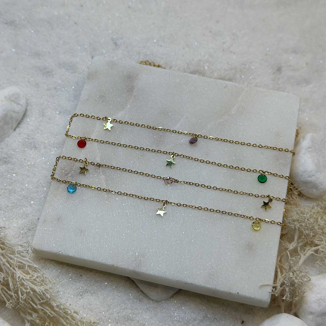 De magnifiques chaînes de taille en or "deli" sont au centre de l'image, posées sur une plaque de marbre. Il y a au total 4 chaînes de taille en or, mais elles possèdent des charms différents : il a des étoiles en or ainsi que des pierres de différentes couleurs (bleu, vert, violet, rouge). En arrière plan se trouve du sable blanc, des cailloux blancs et des algues.