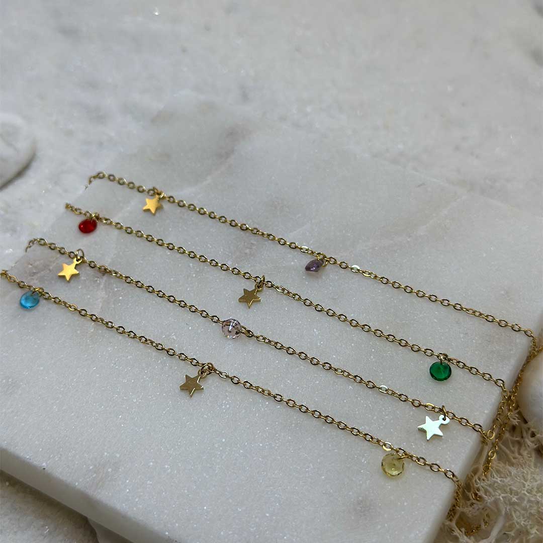 De magnifiques chaînes de taille en or "deli" sont au centre de l'image en gros plan, posées sur une plaque de marbre. Il y a au total 4 chaînes de taille en or, mais elles possèdent des charms différents : il a des étoiles en or ainsi que des pierres de différentes couleurs (bleu, vert, violet, rouge). En arrière plan se trouve du sable blanc.