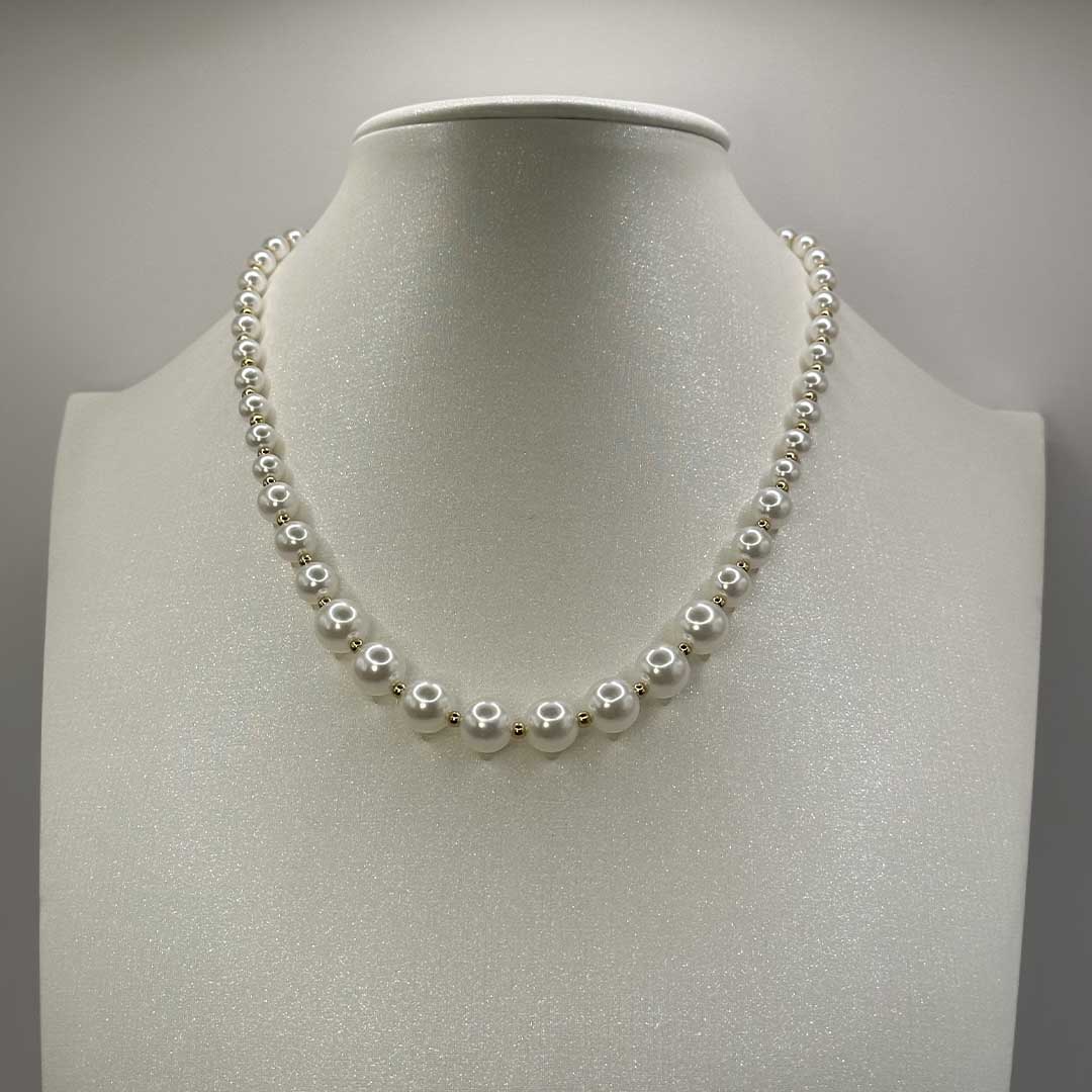 Ce magnifique bijou "Delia" en acier inoxydable est un collier de perles blanches et dorées. Il est posé sur une buste pailleté devant un fond blanc.