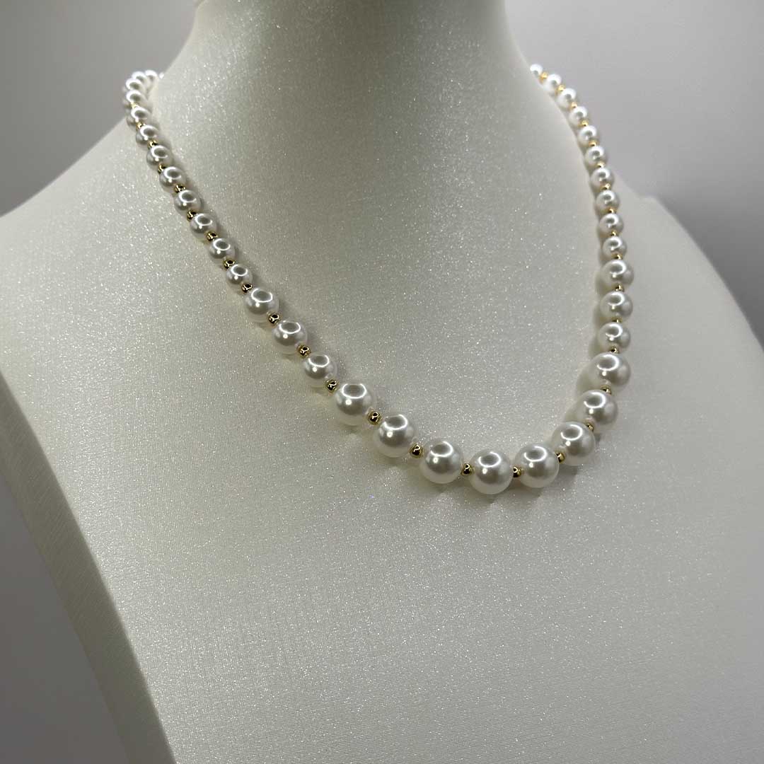 Ce magnifique bijou "Delia" en acier inoxydable est un collier de perles blanches et dorées. Il est posé sur une buste pailleté devant un fond blanc.