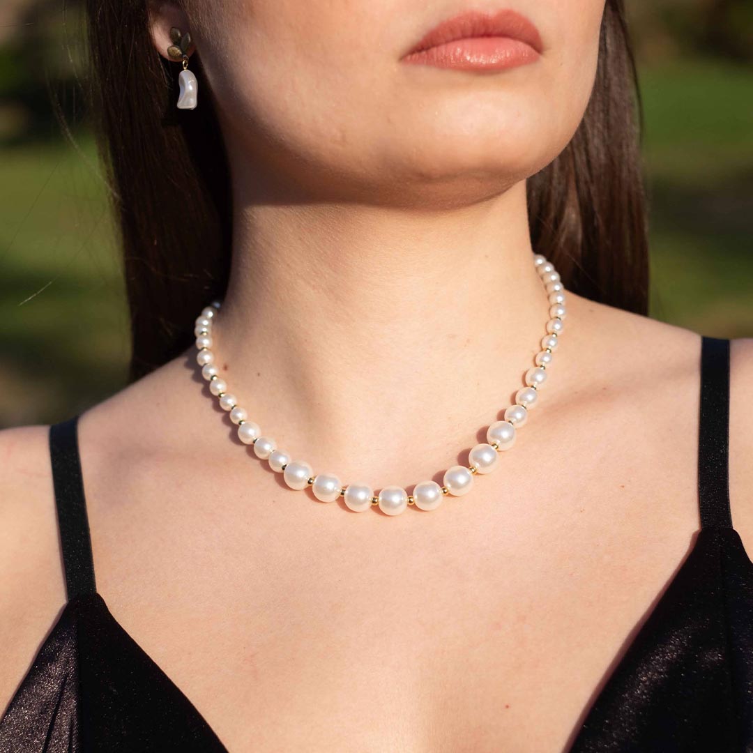 Photo d'un cou et d'un bas de visage montrant ce magnifique bijou "delia" en acier inoxydable qui est un collier de perles blanches nacrées. Il est aussi de couleur doré / or. Les perles font un dégradé de taille. En arrière plan nous pouvons voir les cheveux bruns de la modèle ainsi de ses lèvres.