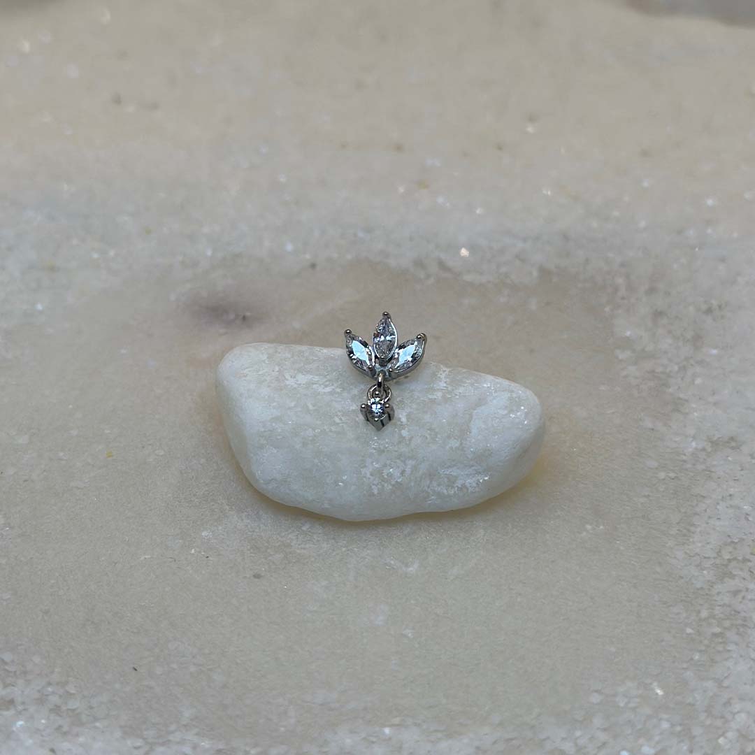 Un très beau piercing en argent "diane" est au centre de l'image, posé sur un caillou blanc. Le bijoux en acier inoxydable est vu de face. Du sable blanc se trouve tout autour.
