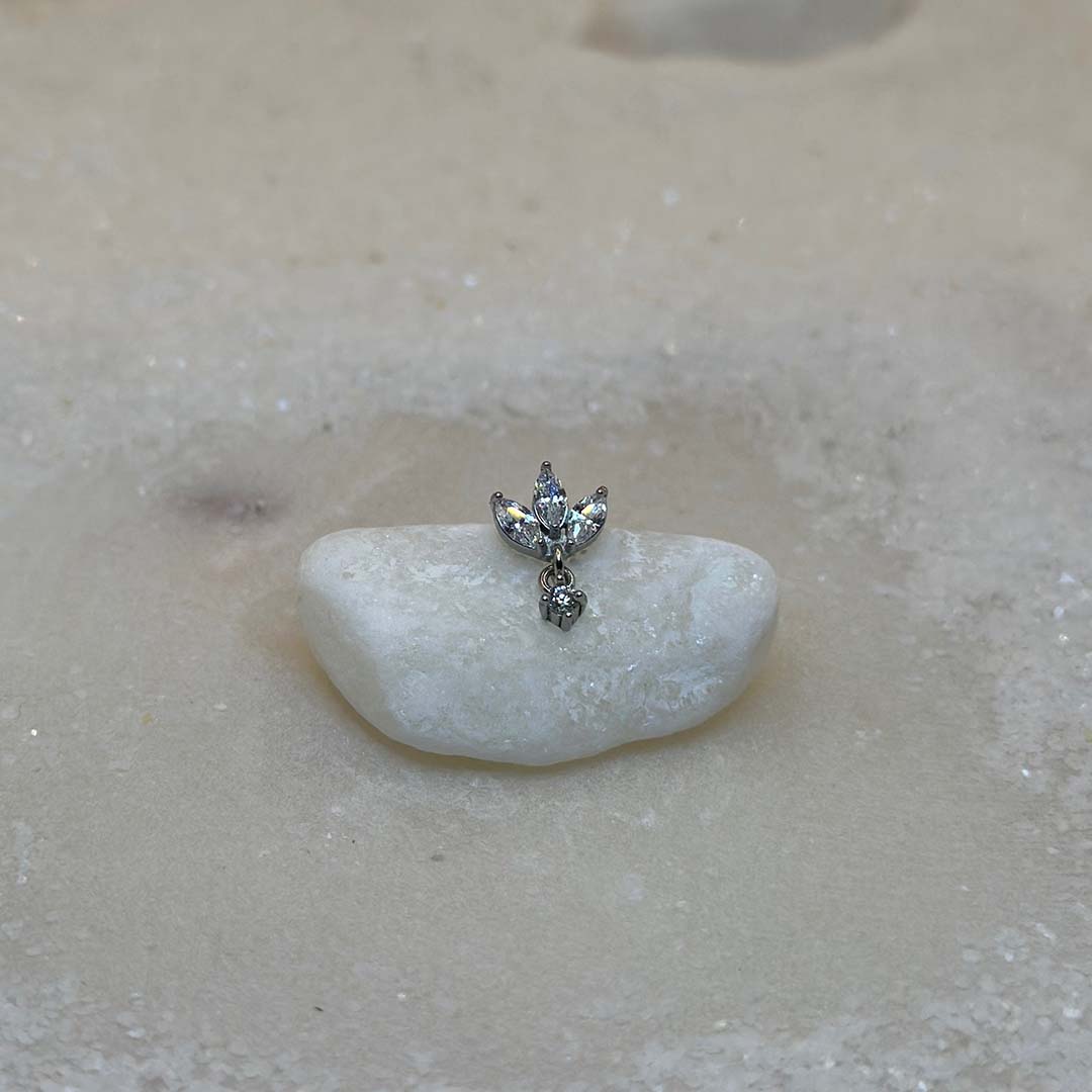 Un très beau piercing en argent "diane" est au centre de l'image, posé sur un caillou blanc. Le bijoux en acier inoxydable est vu de 3/4. Du sable blanc se trouve tout autour.