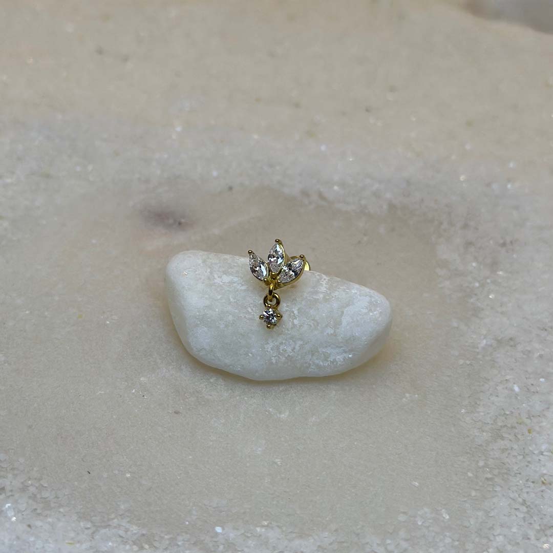 Un très beau piercing en or "diane" est au centre de l'image, posé sur un caillou blanc. Le bijoux en acier inoxydable est vu de 3/4. Du sable blanc se trouve tout autour.