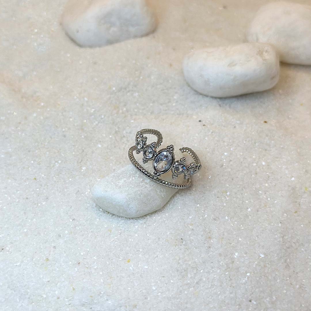 Une très jolie bague en argent "doli" est au centre de l'image, posée sur un petit cailloux blanc. Le bijoux est en acier inoxydable, vu de 3/4, et ressemble à une couronne. Autour se trouvent d'autres cailloux et du sable blanc.