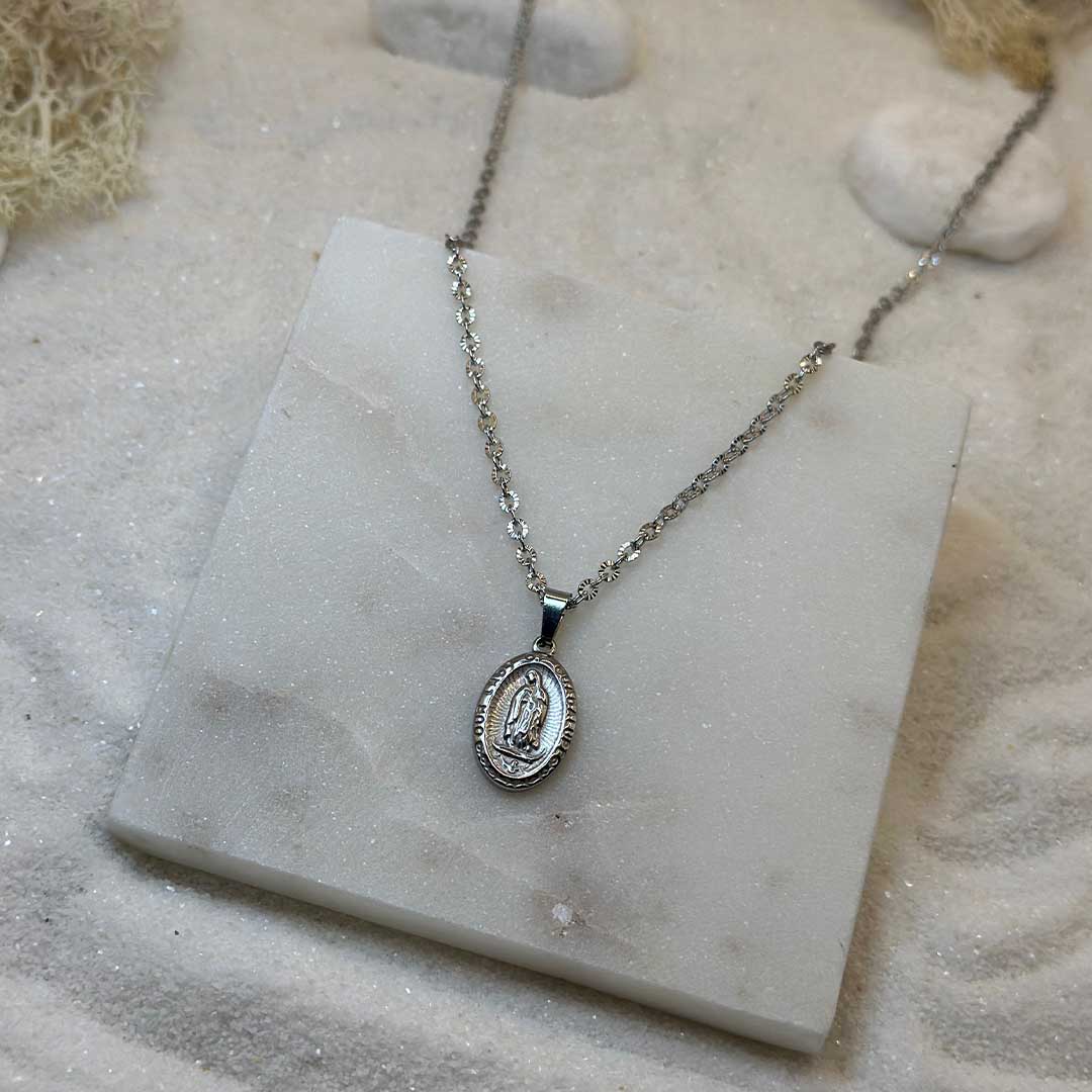 Un très jolie collier en argent "eden" est au centre de l'image, posé sur une plaque de marbre. Le bijoux en acier inoxydable ressemble à un collier chrétien, avec un pendentif et d'un saint. Autour se trouve du sable blanc.