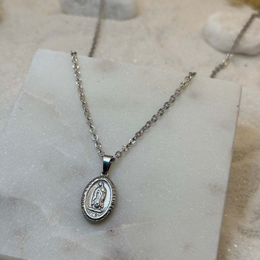 Un très jolie collier en argent "eden" est au centre de l'image en gros plan, posé sur une plaque de marbre. Le bijoux en acier inoxydable ressemble à un collier chrétien, avec un pendentif et d'un saint. Autour se trouve du sable blanc.