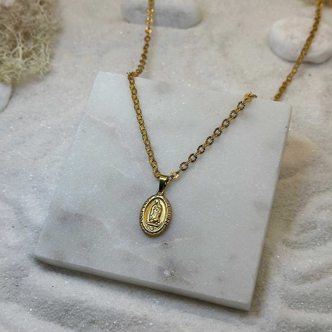 Un très jolie collier en or "eden" est au centre de l'image, posé sur une plaque de marbre. Le bijoux en acier inoxydable ressemble à un collier chrétien, avec un pendentif et d'un saint. Autour se trouve du sable blanc.