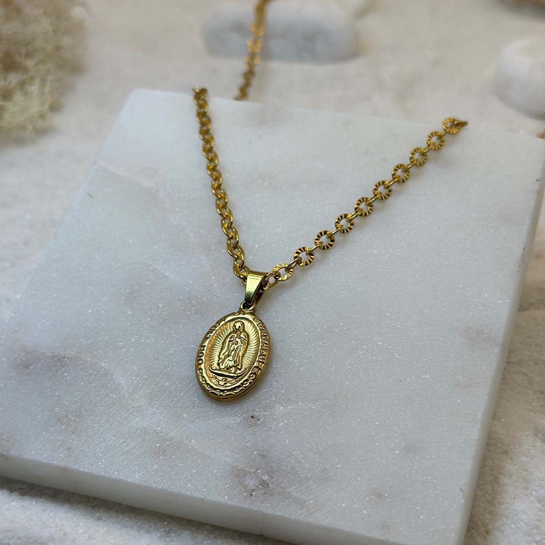 Un très jolie collier en or "eden" est au centre de l'image en gros plan, posé sur une plaque de marbre. Le bijoux en acier inoxydable ressemble à un collier chrétien, avec un pendentif et d'un saint. Autour se trouve du sable blanc.
