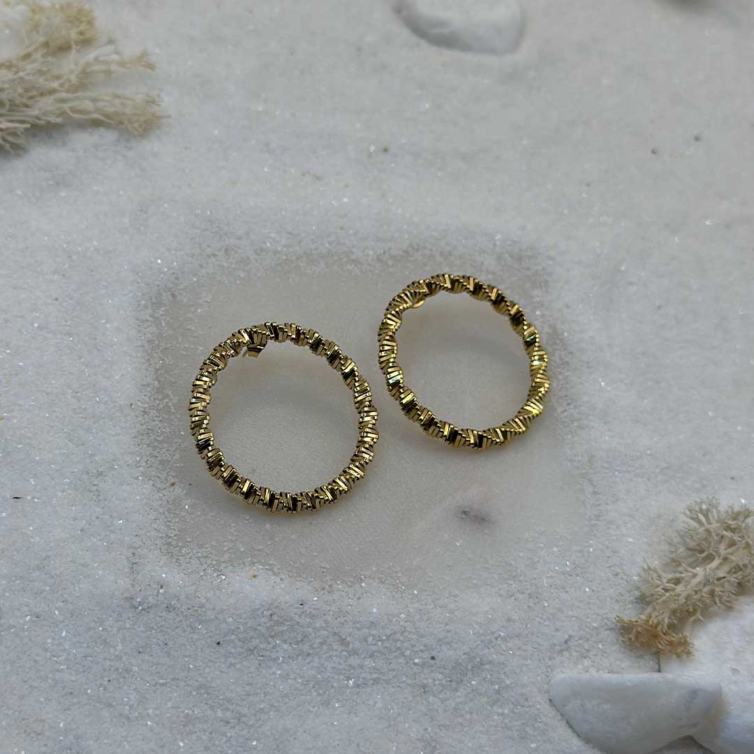Une très jolie paire de boucles d'oreille en or "emy" est au centre de l'image. Les bijoux sont en acier inoxydable, et posés sur du sable blanc. Autour se trouvent des cailloux et des algues.