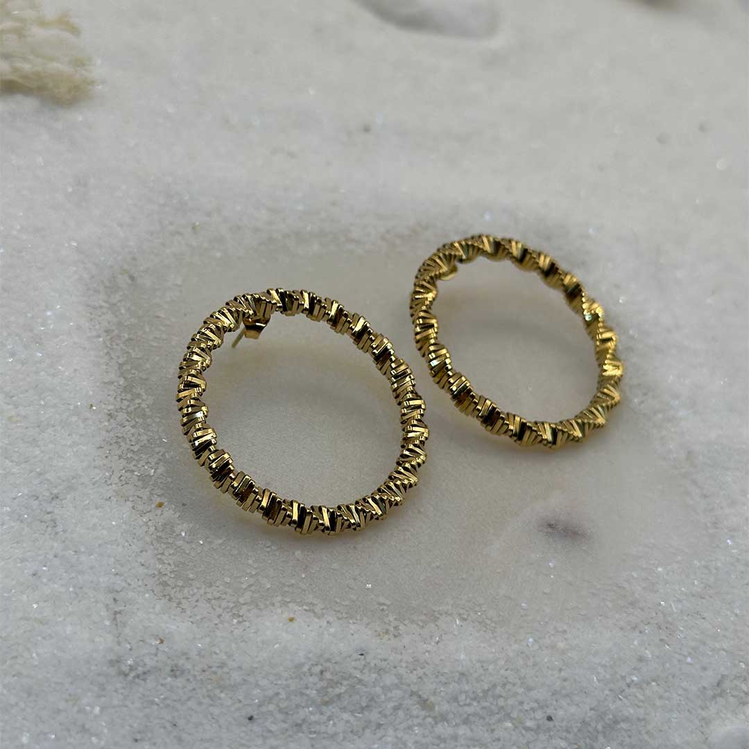 Une très jolie paire de boucles d'oreille en or "emy" est au centre de l'image. Les bijoux sont en acier inoxydable, et posés sur du sable blanc en gros plan. Autour se trouvent des cailloux et des algues.