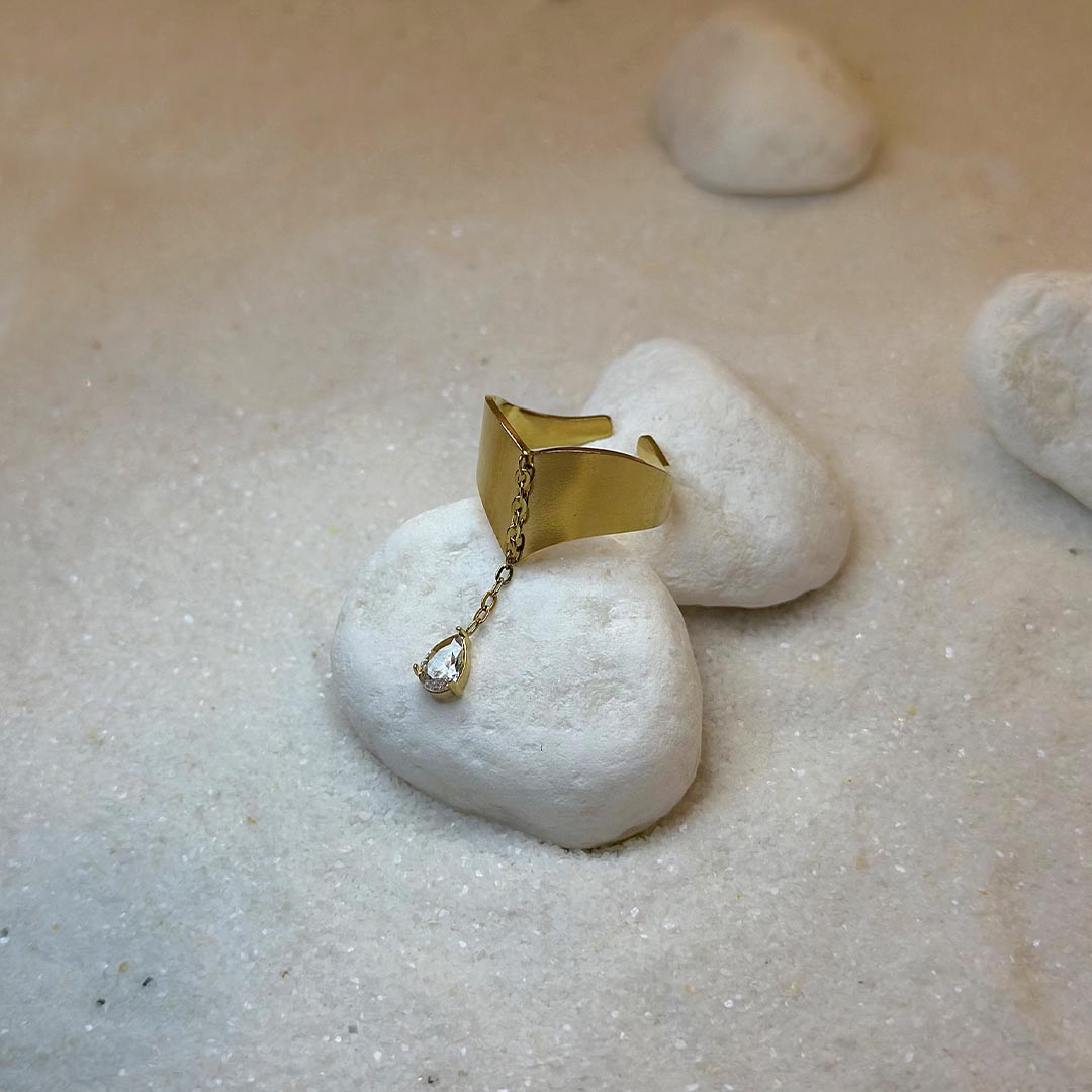 Une très jollie bague en or "enea" est au centre de l'image, posée sur un cailloux blanc. Le bijoux en acier inoxydable est en deux parties : la première est une base en or, en forme de flèche. La seconde est un strass, relié par une chaîne en or à la base. Autour se trouve du sable blanc.