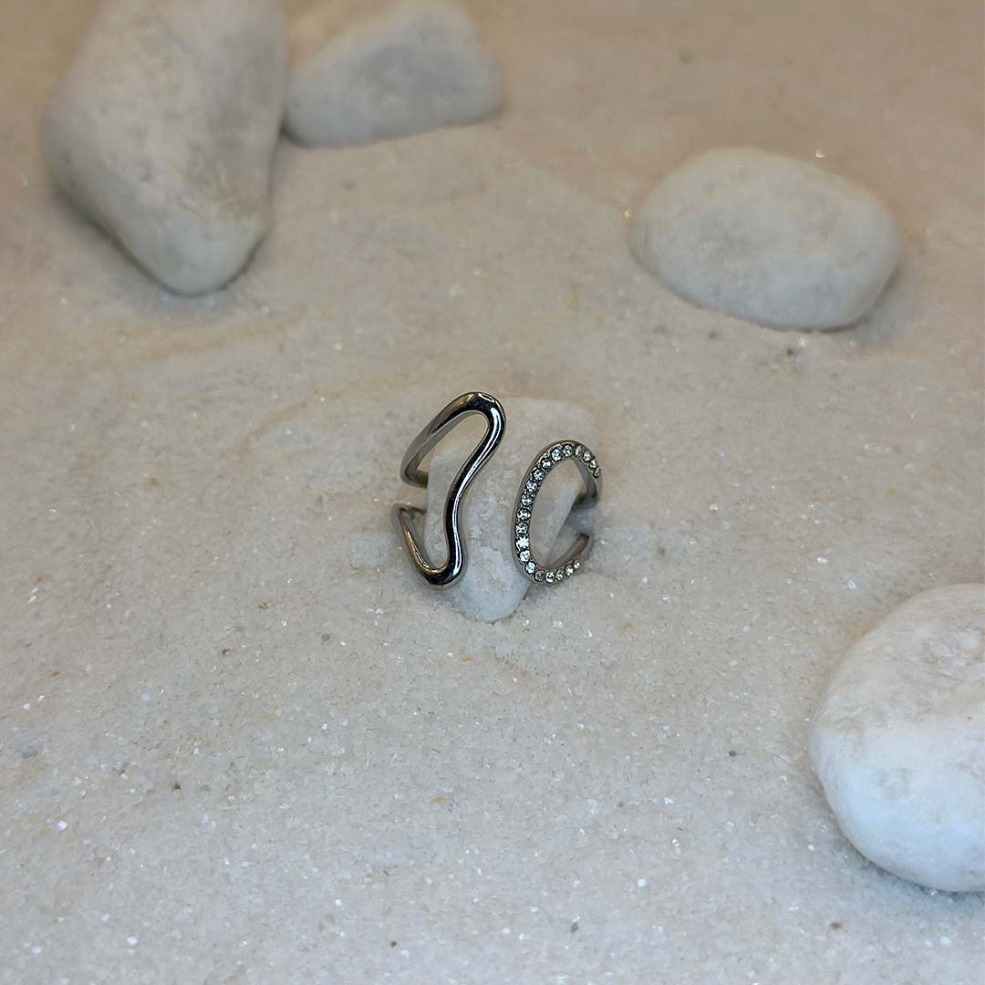 Une magnifique bague en argent "era" est au centre de l'image, posées sur du sable et blanc et entourant un caillou blanc. Le bijoux en acier inoxydable possède des strass sur sa partie droite. Autour se trouvent d'autres cailloux et du sable.