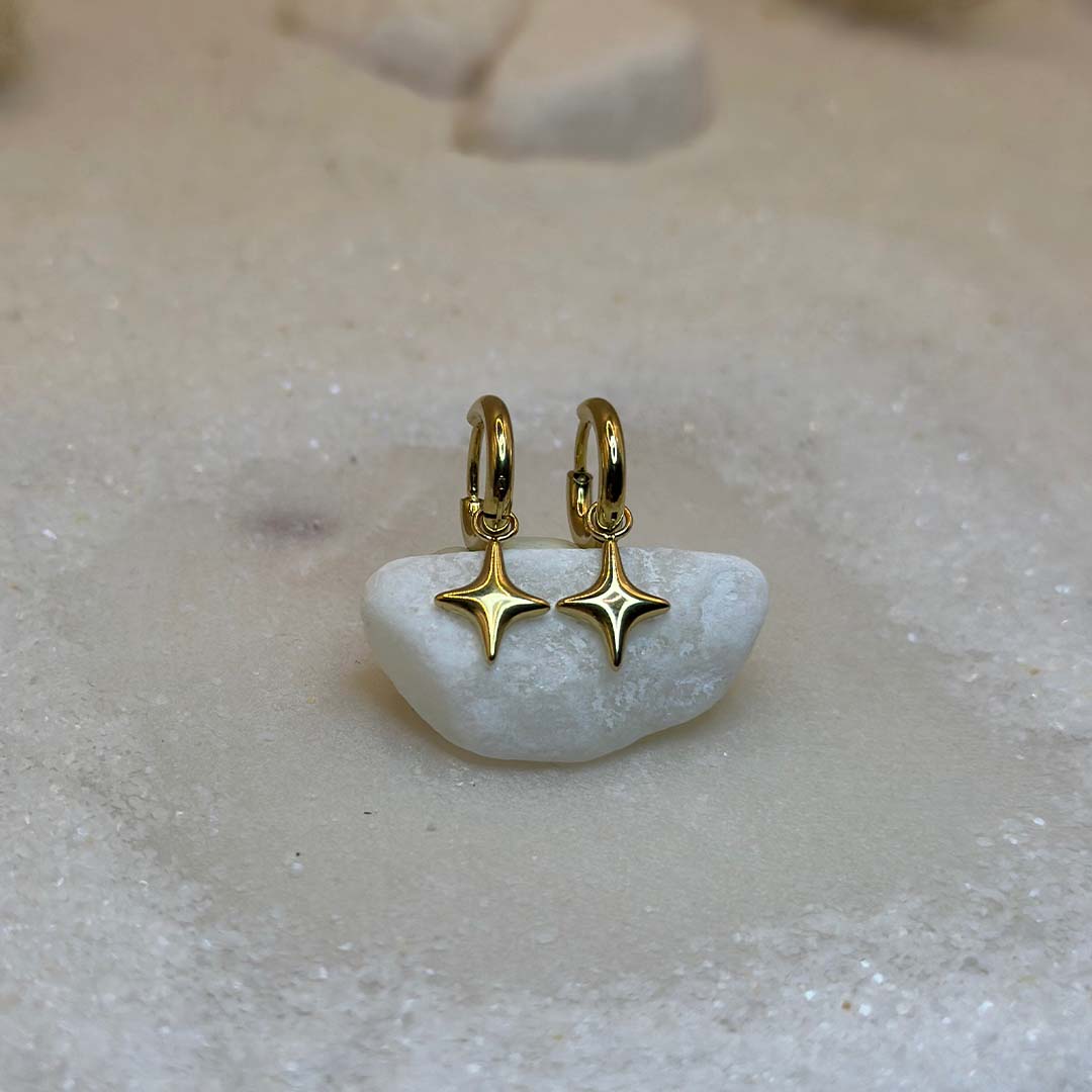 Deux très belles boucles d'oreilles en or "estella", sont au centre de l'image, posées sur un cailloux blanc et vu de face. Ce sont des bijoux en acier inoxydable en forme d'étoiles. Tout autour se trouve du sable blanc.