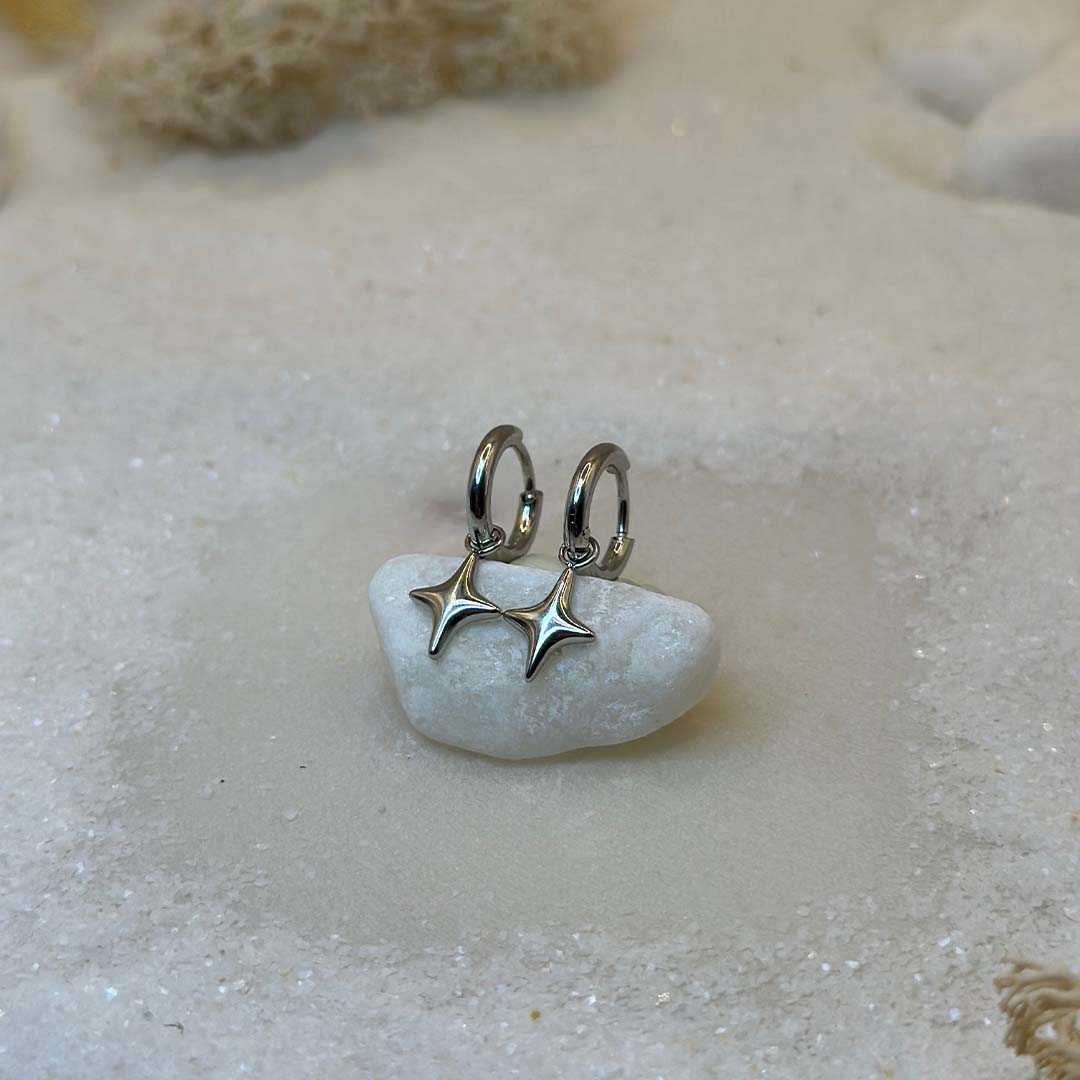 Deux très belles boucles d'oreilles en argent "estella", sont au centre de l'image, posées sur un cailloux blanc et vu de 3/4. Ce sont des bijoux en acier inoxydable en forme d'étoiles. Tout autour se trouve du sable blanc.