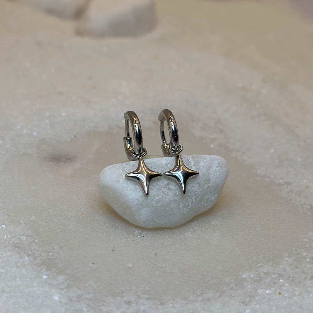 Deux très belles boucles d'oreilles en argent "estella", sont au centre de l'image, posées sur un cailloux blanc et vu de face Ce sont des bijoux en acier inoxydable en forme d'étoiles. Tout autour se trouve du sable blanc.