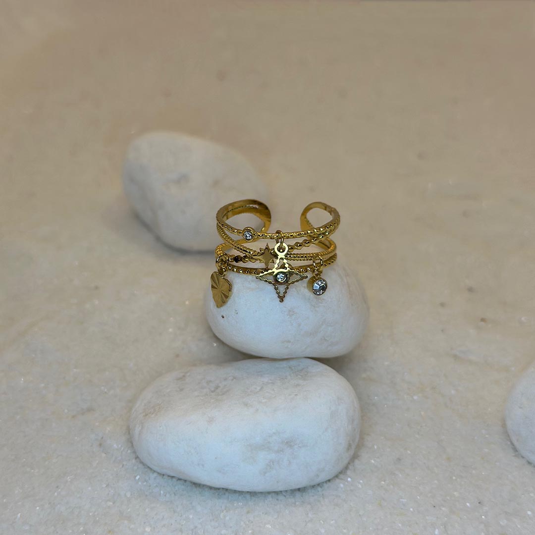 Une jolie bague en or "eve" est au centre de l'image, joliment posée sur un caillou blanc. Le bijoux en acier inoxydable est composé de plusieurs détails : des charms : une étoiles, un strass, une feuille. Ce sont des plusieurs bagues, réunies en une seule. Tout autour se trouve du sable blanc et d'autres cailloux.