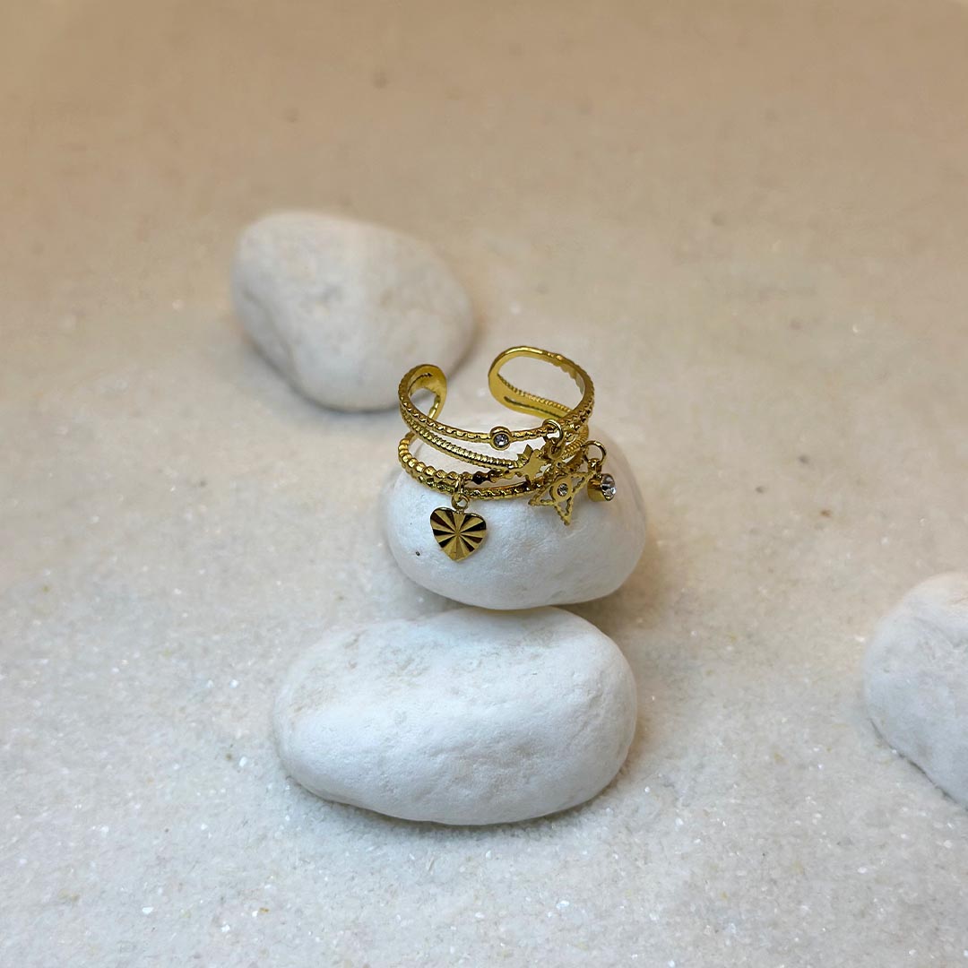 Une jolie bague en or "eve" est au centre de l'image, joliment posée sur un caillou blanc et vu d'en haut. Le bijoux en acier inoxydable est composé de plusieurs détails : des charms : une étoiles, un strass, une feuille. Ce sont des plusieurs bagues, réunies en une seule. Tout autour se trouve du sable blanc et d'autres cailloux.