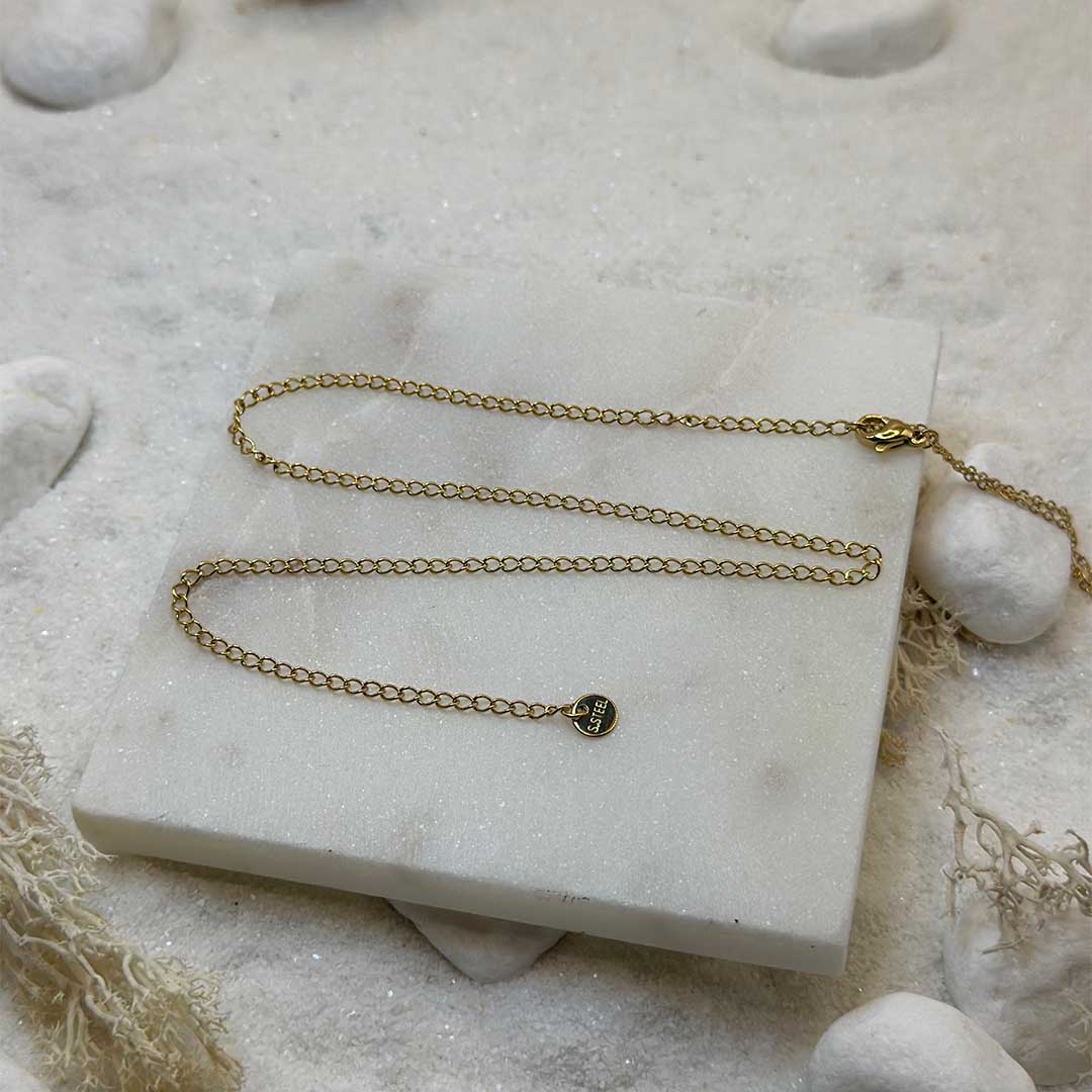 Une magnifique extension de chaîne de ventre en or est au centre de l'image, magnifiquement posée sur une plaque de marbre. Le bijoux en acier inoxydable est composé de maillons en or. Autour se trouve du sable blanc, des cailloux et des algues.