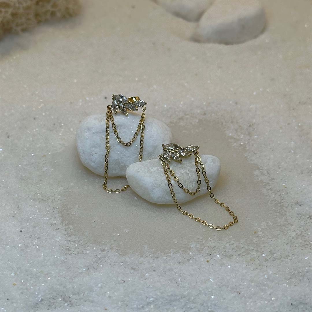 Une magnifique paire de boucle d'oreilles en or "Ezea" est au centre de l'image, vu de face, posé sur une grosse plaque de marbre. Le bijoux en acier inoxydable est composée de feuille en strass et de deux petites chaines. Autour se trouve du sable blanc.