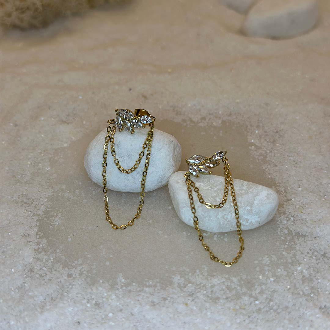 Une magnifique paire de boucle d'oreilles en or "Ezea" est au centre de l'image, vu de face, posé sur une grosse plaque de marbre. Le bijoux en acier inoxydable est composée de feuille en strass et de deux petites chaines. Autour se trouve du sable blanc.