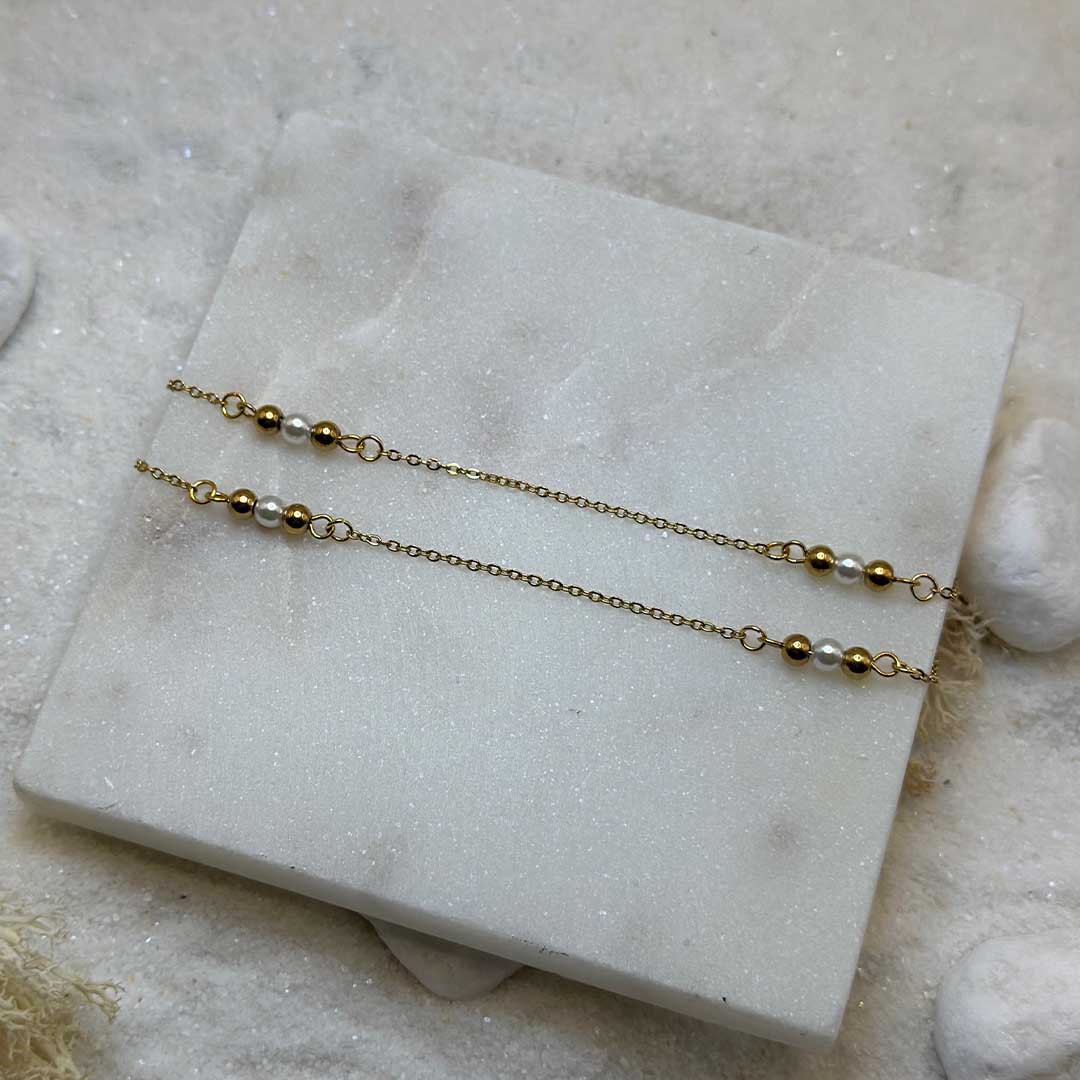 Une très belle chaîne de taille en or "felicie" est au centre de l'image, vu penchée. Le bijoux en acier inoxydable possède des perles en or et perles basiques, les maillons sont en or. Les deux chaînes sont posées sur une plaque de marbre. Tout autour se trouve du sable blanc, des cailloux et des algues.