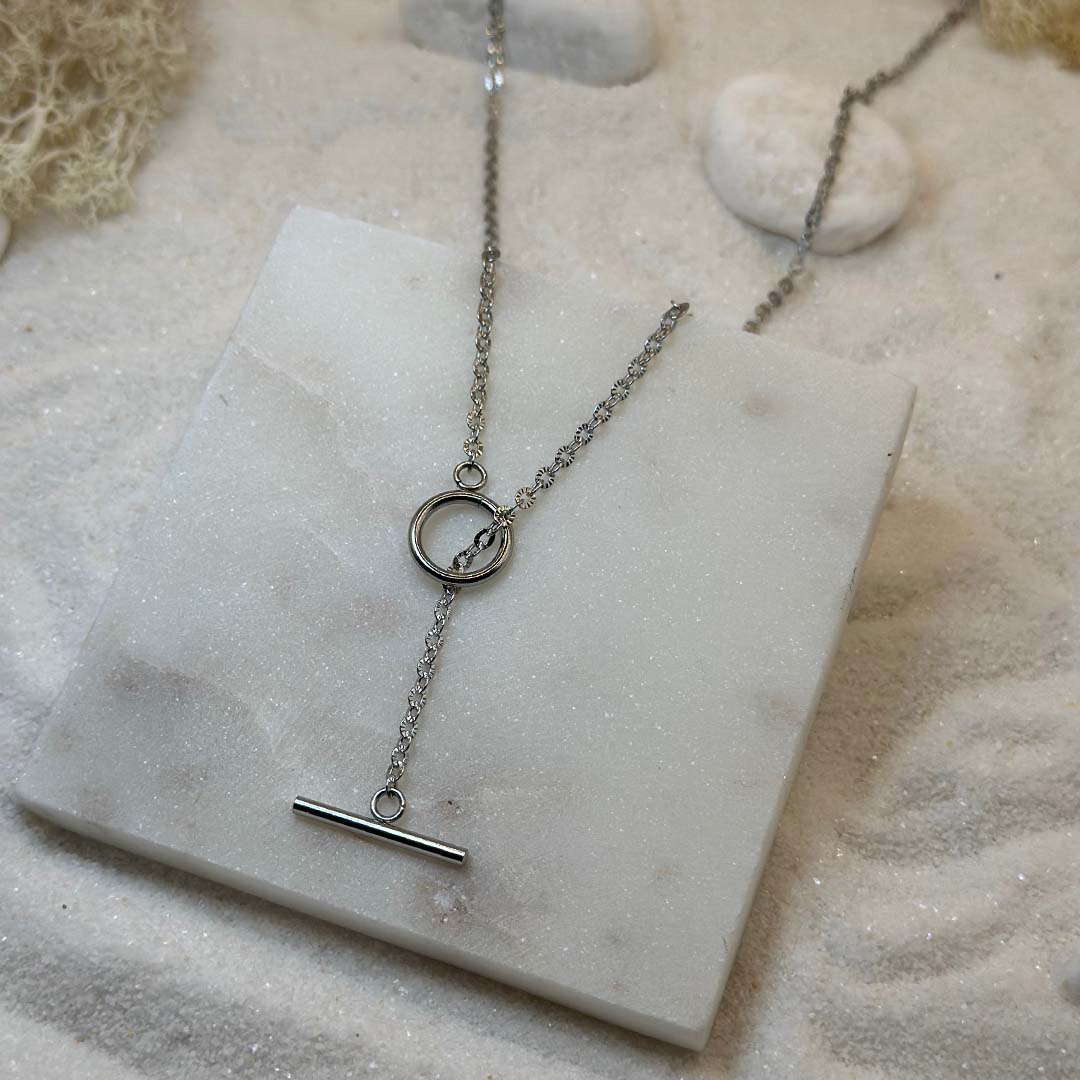 Un magnifique collier en argent "fenicie" est au centre de l'image, posé sur une plaque en marbre. Le bijoux en acier inoxydable est composé d'une chaîne en forme de maillons étoilés, ainsi que d'un cercle et d'une barre lisses. En fond se trouve du sable blanc.