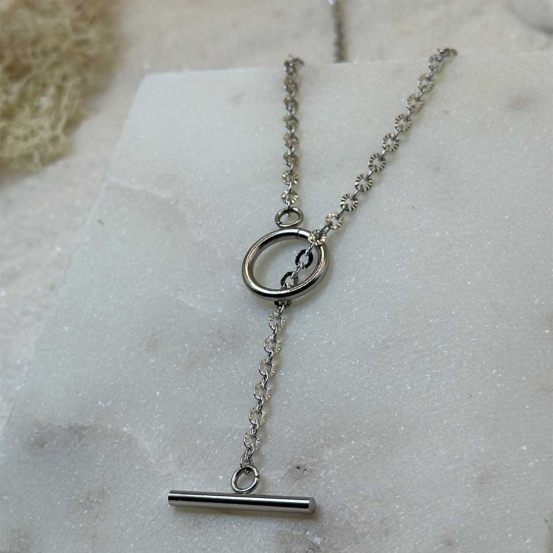 Un magnifique collier en argent "fenicie" est au centre de l'image, posé sur une plaque en marbre en gros plan. Le bijoux en acier inoxydable est composé d'une chaîne en forme de maillons étoilés, ainsi que d'un cercle et d'une barre lisses. En fond se trouve du sable blanc.