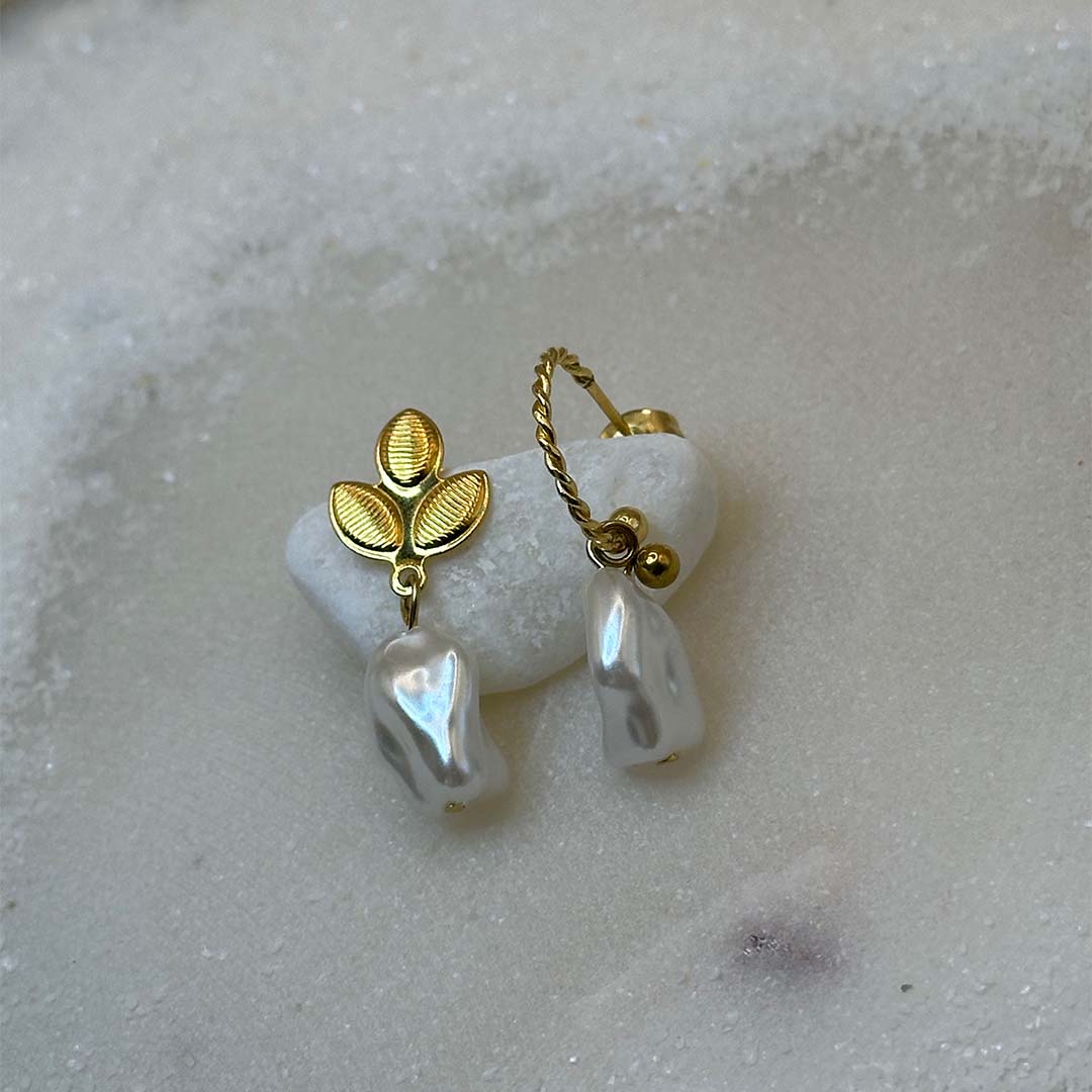 De très belles boucles d'oreilles en or avec des perles "fidji", sont au centre de l'image, en gros plan, et posées sur un caillou blanc. Les bijoux en acier inoxydable sont vu de 3/4. Tout autour se trouve du sable blanc.