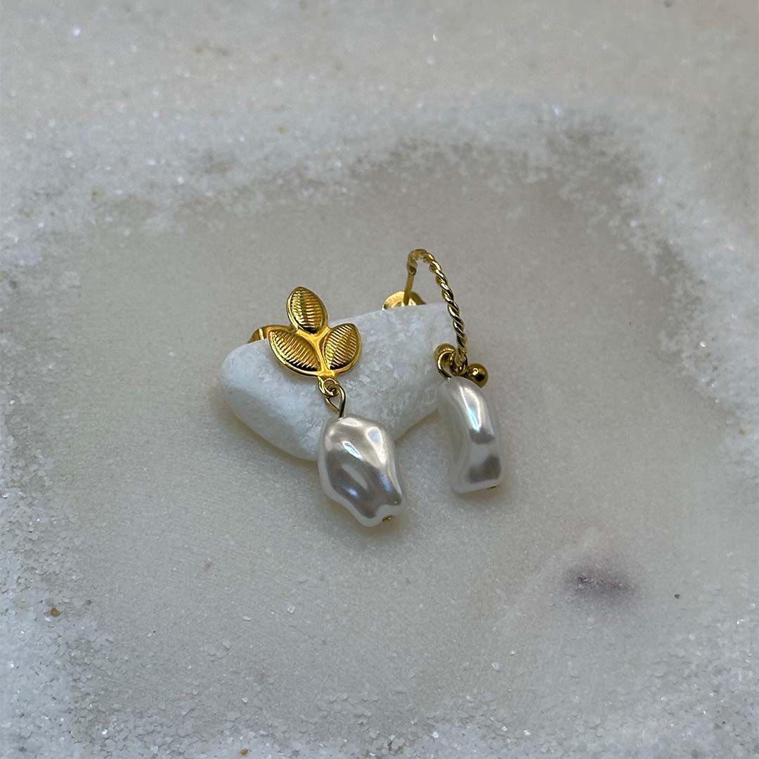 De très belles boucles d'oreilles en or avec des perles "fidji", sont au centre de l'image, en gros plan, et posées sur un caillou blanc. Les bijoux en acier inoxydable sont vu de 3/4. Tout autour se trouve du sable blanc.