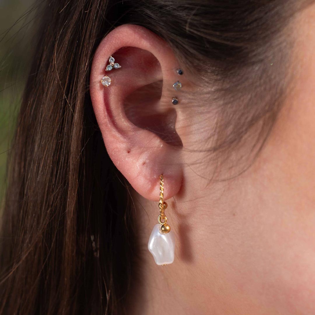 Photo d'une oreille prise de près pour montrer ce magnifique bijou "fidji" en acier inoxydable qui est une boucle d'oreille avec un anneau torsadé auquel est accroché une petite boule de couleur or et une perle nacrée blanche. . En arrière plan nous pouvons voir les cheveux bruns de la modèle ainsi de des piercings.