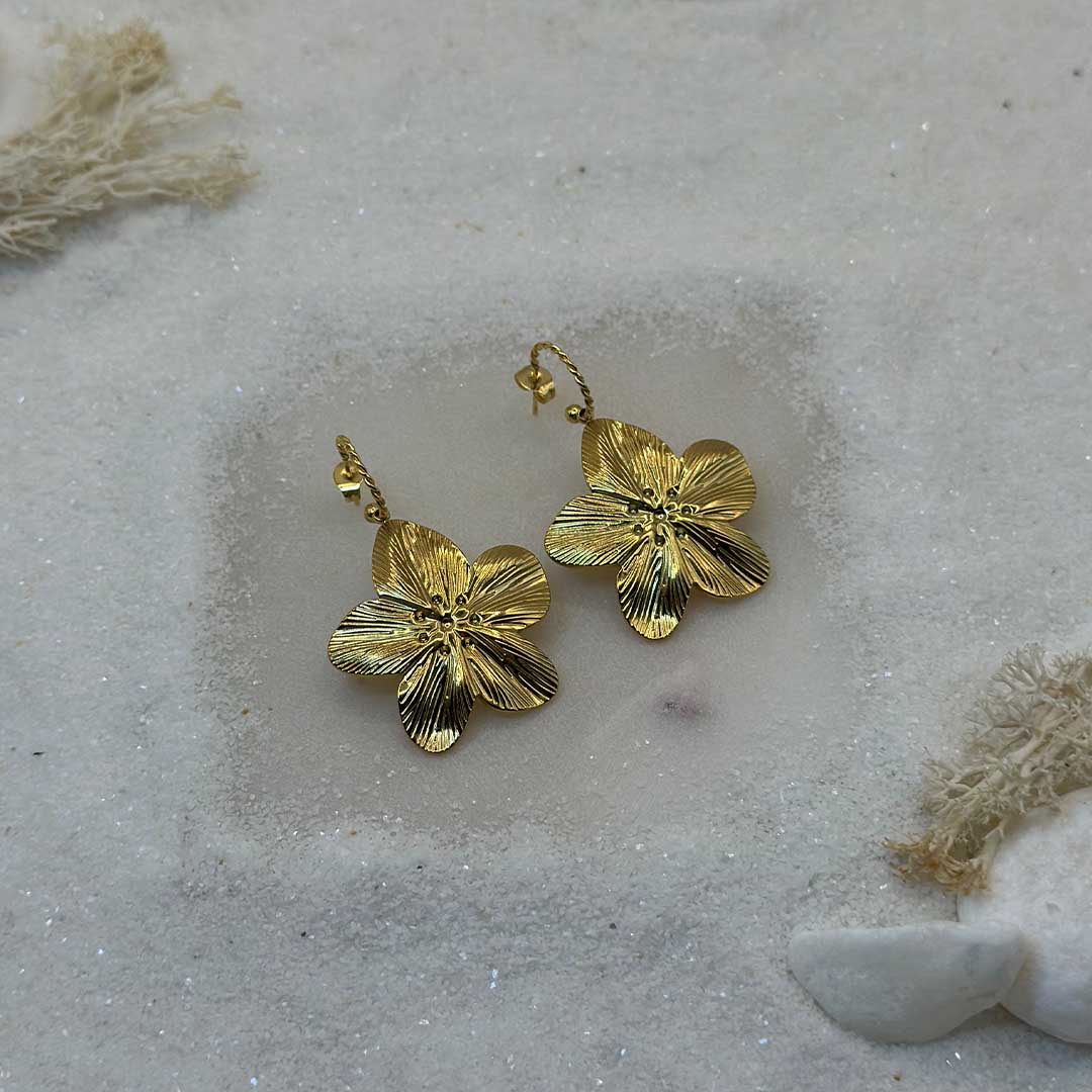 De très belles boucles d'oreille en or "flora" sont au centre de l'image, posées sur du sable blanc. Les bijoux en acier inoxydable sont en forme de fleurs, et vu d'en haut. Tout autour se trouve du sable blanc.