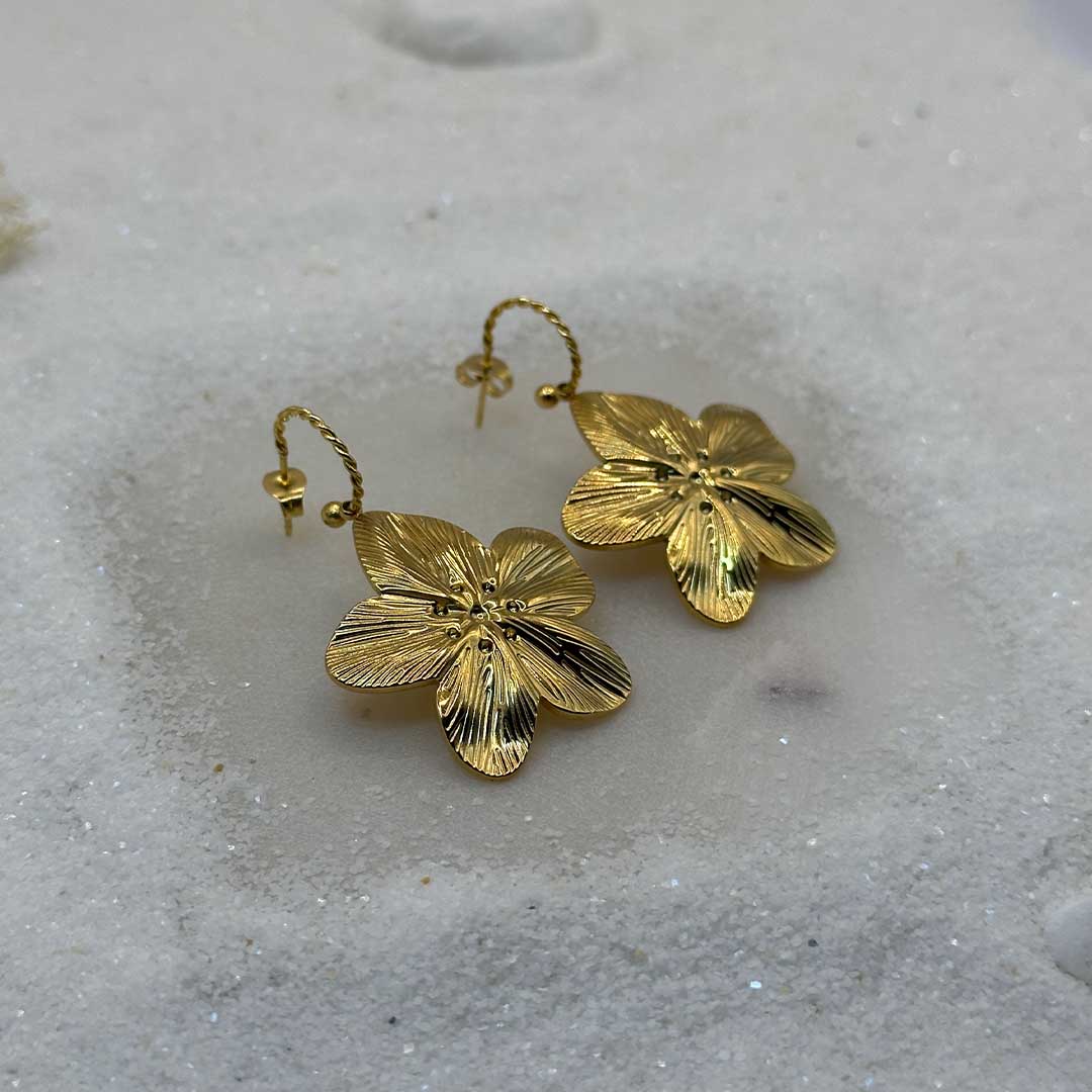 De très belles boucles d'oreille en or "flora" sont au centre de l'image, posées sur du sable blanc. Les bijoux en acier inoxydable sont en forme de fleurs, et vu de 3/4 en gros plan. Tout autour se trouve du sable blanc.