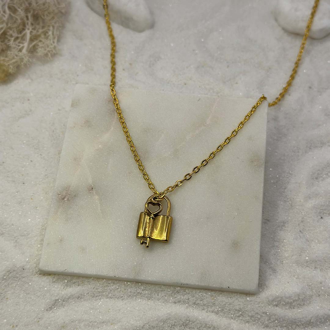 Un magnifique collier en or "Ginie" est au centre de l'image, vu de face, posé sur une grosse plaque de marbre. Le bijoux en acier inoxydable est composée d'une chaine en or, de pendentifs en forme de cadenas et une clef. Autour se trouve du sable blanc.