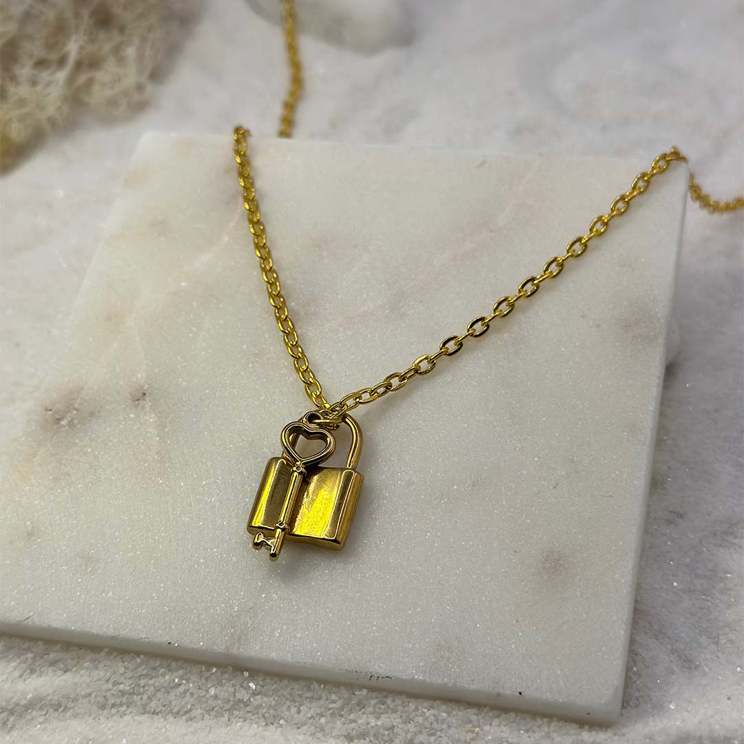 Un magnifique collier en or "Ginie" est au centre de l'image, vu de face, posé sur une grosse plaque de marbre. Le bijoux en acier inoxydable est composée d'une chaine en or, de pendentifs en forme de cadenas et une clef. Autour se trouve du sable blanc.
